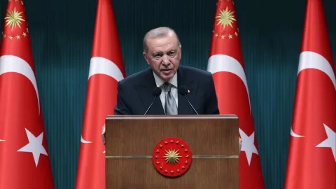Cumhurbaşkanı Erdoğan konuşurken, mikrofonlar ve Türk bayrağı arka planda görünüyor
