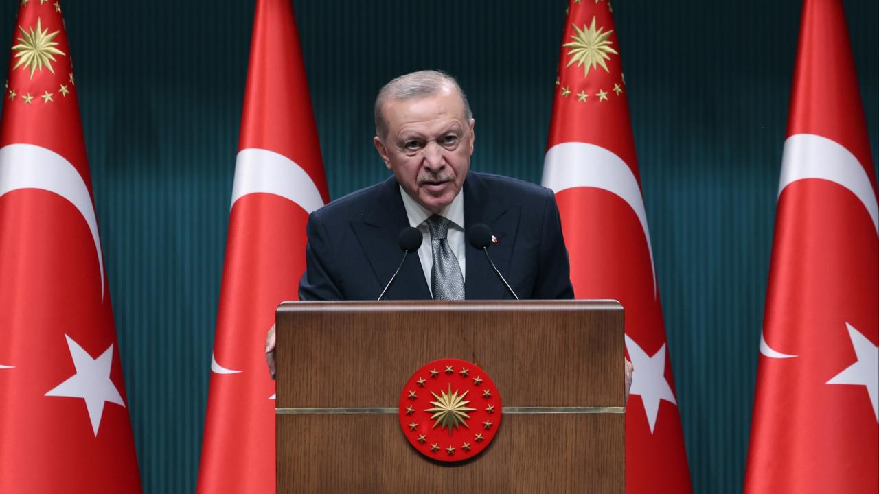 Cumhurbaşkanı Erdoğan konuşurken, mikrofonlar ve Türk bayrağı arka planda görünüyor