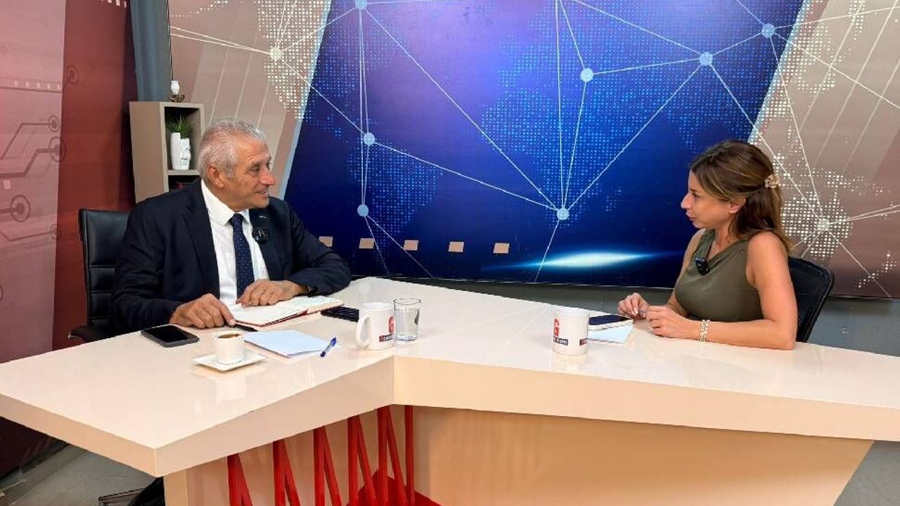 Taçoy konuşurken resmi; arka planda bayrak ve masa, resmi toplantı ortamı