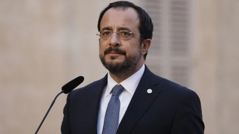 Nikos Hristodulidis konuşurken ciddi bir ifadeyle mikrofon önünde duruyor.
