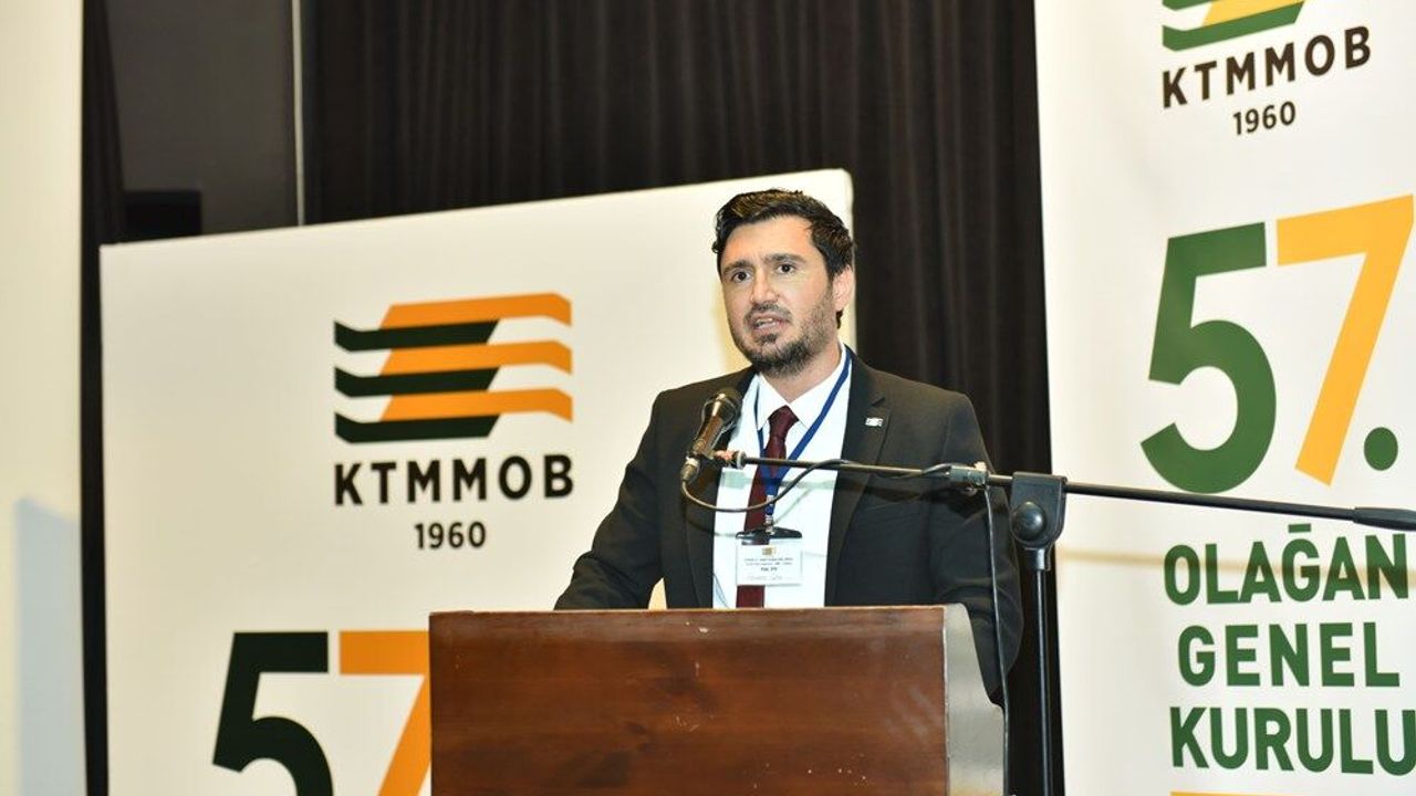 Çelik konuşurken mikrofon önünde duruyor, arka planda toplantı veya kürsü ortamı
