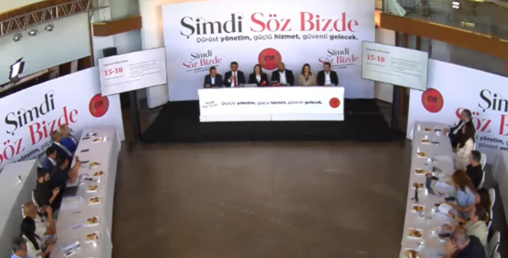 İncirli oturarak mikrofonla konuşuyor; arka planda tanıtım görselleri ve bayraklar görünüyor.