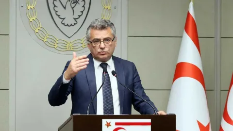 Erhürman konuşurken ceketli, mikrofon ve bayrak arka planda görünüyor olabilir