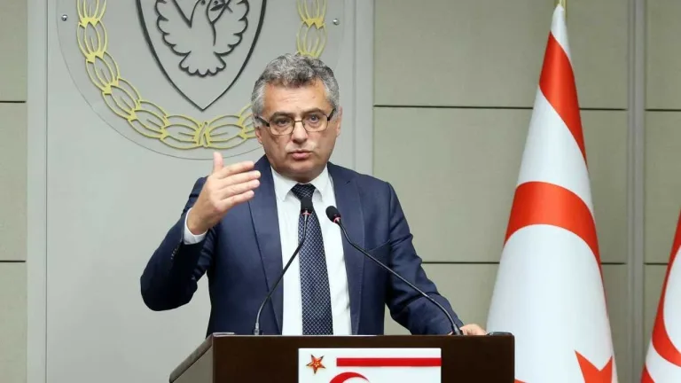 Erhürman konuşurken ceketli, mikrofon ve bayrak arka planda görünüyor olabilir
