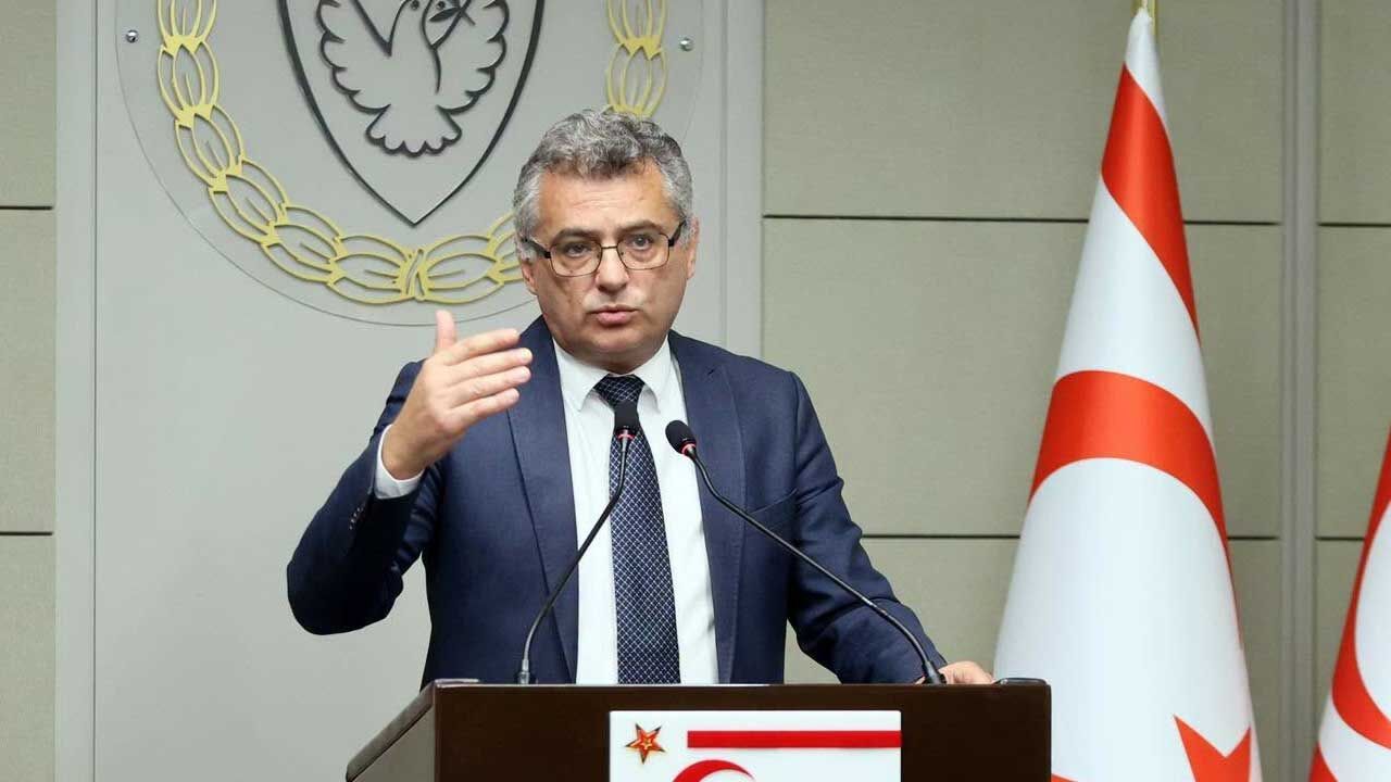 Erhürman konuşurken ceketli, mikrofon ve bayrak arka planda görünüyor olabilir