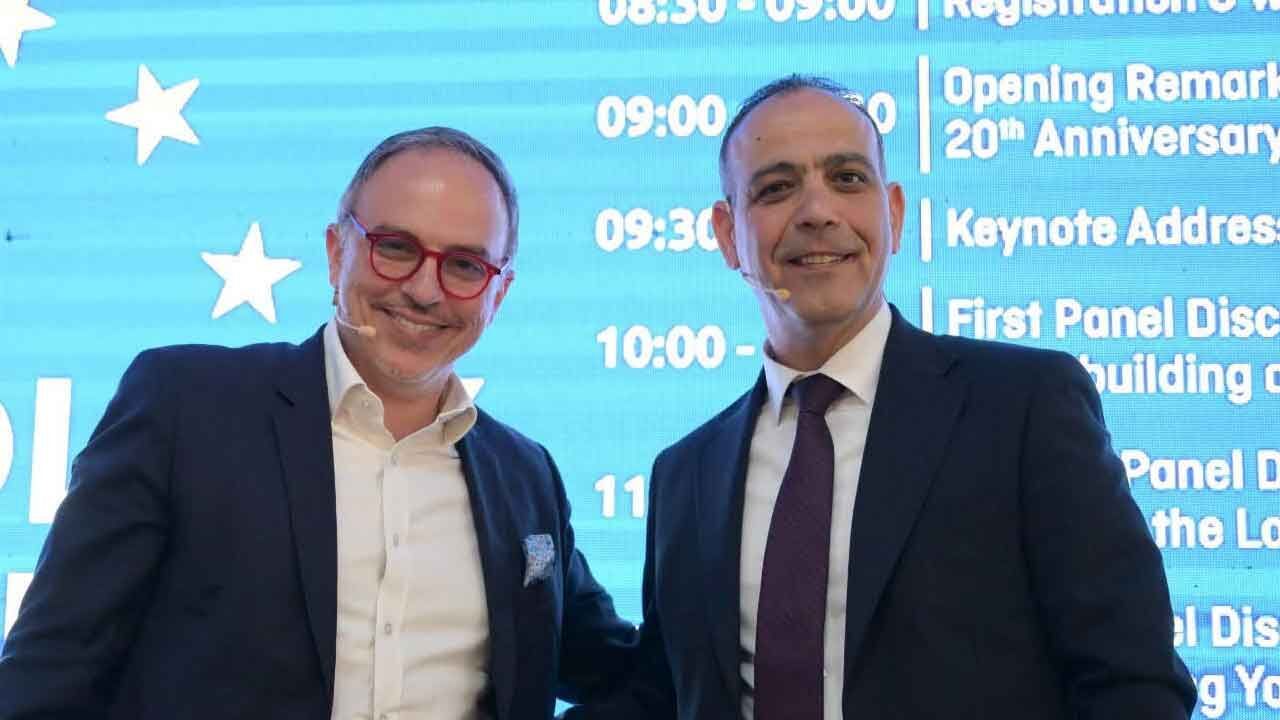 Harmancı konuşma yaparken el hareketiyle vurgu yapıyor, arka planda Türk ve Avrupa Birliği bayrakları görünüyor olabilir