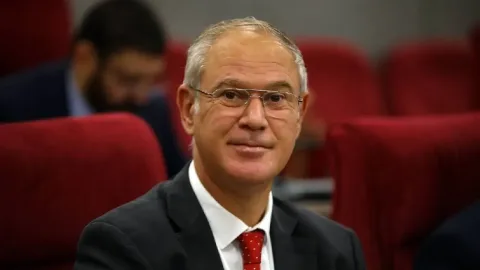 Hasipoğlu konuşuyor; arka planda bir pankart veya salon ortamı görünüyor