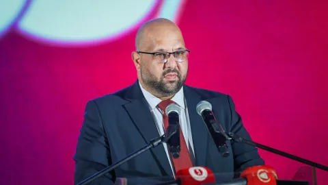 Mehmet Kale konuşuyor; arka planda flu bir iç mekan ve mikrofonlar görünüyor