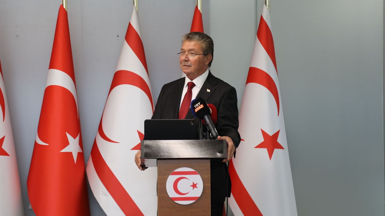 Üstel konuşma yaparken kürsüde duruyor, arka planda etkinlik dekoru ve seyirciler görünüyor