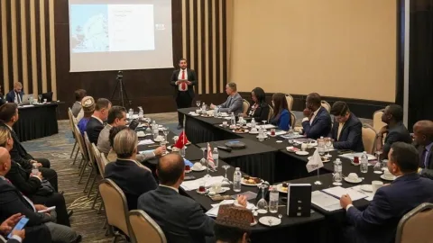 Ataoğlu konuşurken mikrofon önünde, arka planda etkinlik görselleri ve katılımcılar bulunuyor