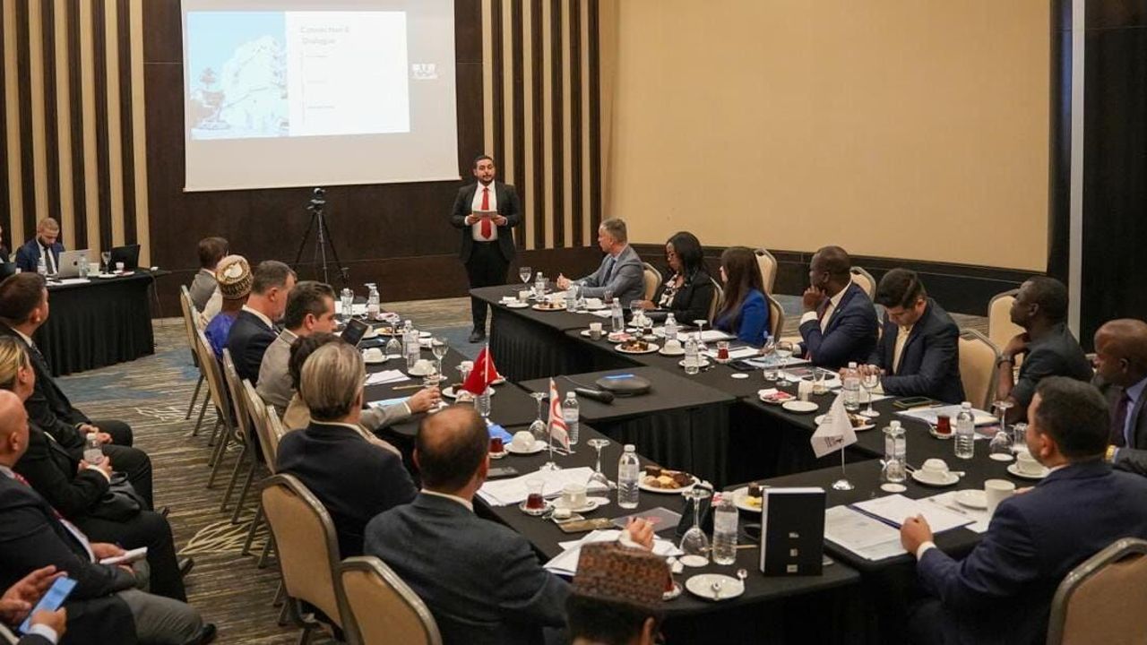 Ataoğlu konuşurken mikrofon önünde, arka planda etkinlik görselleri ve katılımcılar bulunuyor