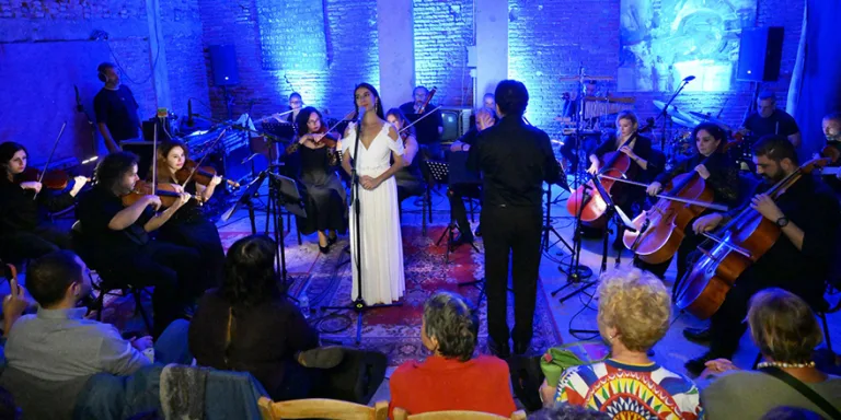 Lefkoşa Belediye Orkestrası ile Gigi sahnede, konser atmosferi ve enstrümanlar görünür.
