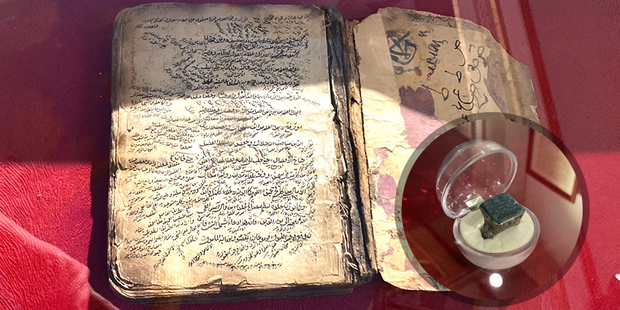 Dört el yazması eser rafta sergileniyor; arka planda müze iç mekânı ve vitrini görülüyor