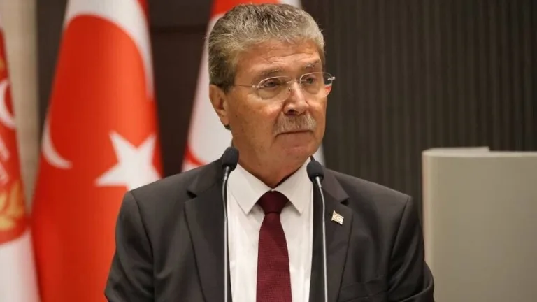 Üstel konuşurken mikrofonla bakıyor, arka planda projelere dair görseller veya katılımcılar olabilir