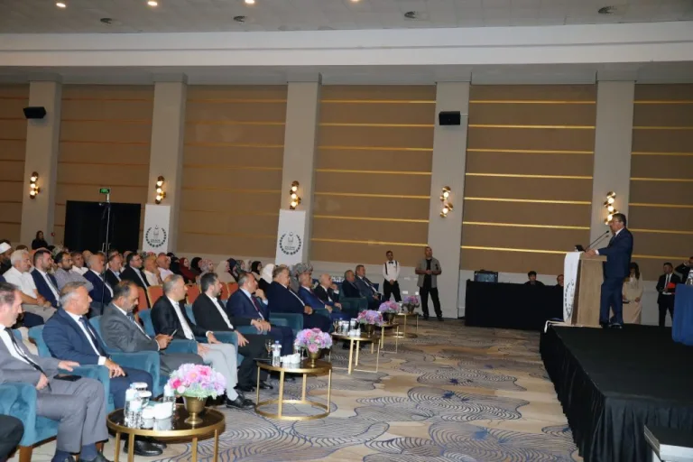 Üstel konuşurken ciddiyetle bakıyor; arka planda bayrak ve gençler görünür gibi