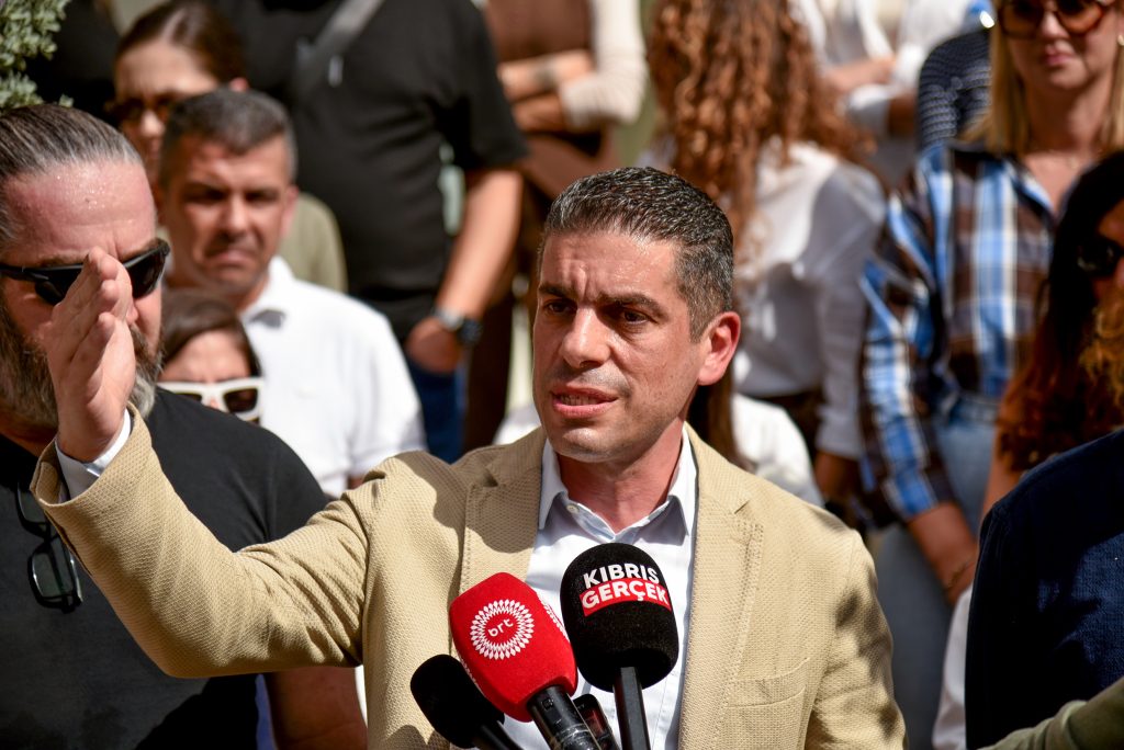 KTAMS Başkanı Yiğit konuşurken ciddi ifadeyle el hareketi yapıyor, arka planda parti ya da sendika simgeleri görünür.