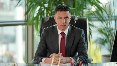 Amcaoğlu konuşurken mikrofonla bakıyor; arka planda kutlama ya da ödül töreni atmosferi