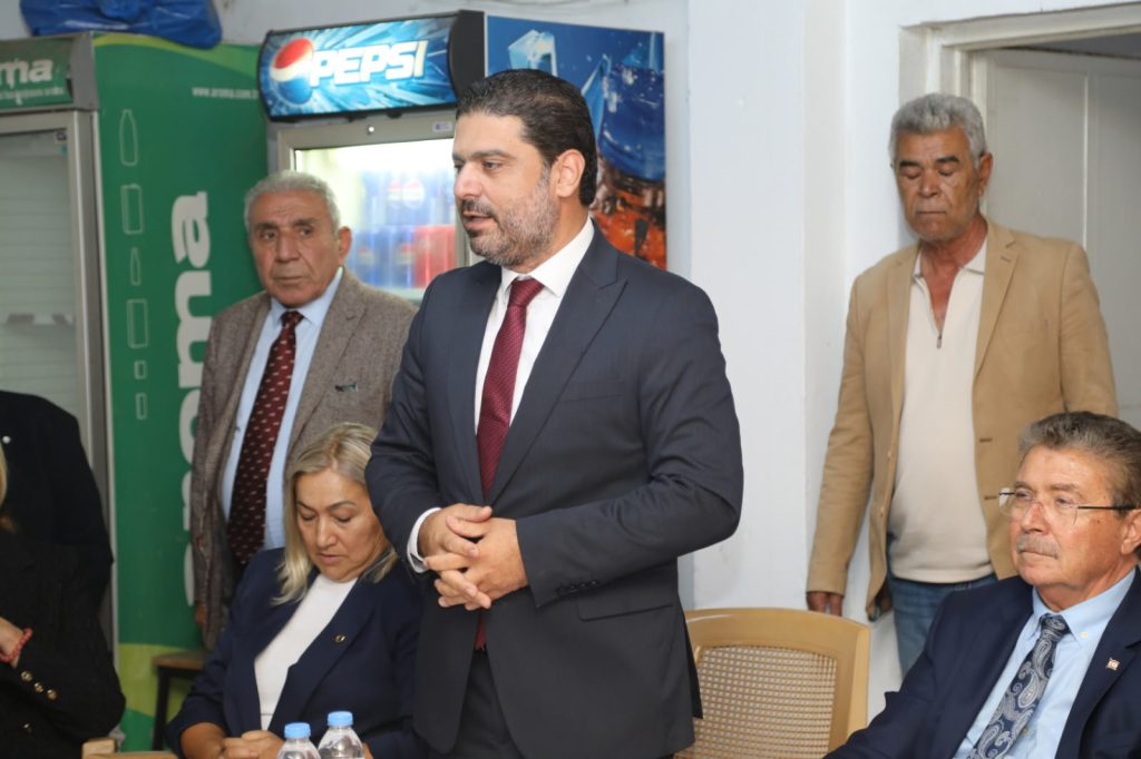 Üstel konuşma yaparken kürsüde mikrofonlarla görülüyor, arka planda parti görselleri.