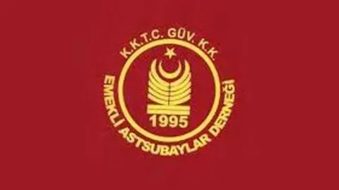 Ömürlü konuşurken arkada Türk ve KKTC bayrakları görünür; ciddî ifadeyle dinleyenler var
