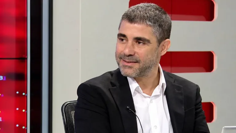 Aktunç konuşurken elleriyle vurgulama yapıyor; arkada Lefkoşa'ya dair şehir unsurları sugeren sahne