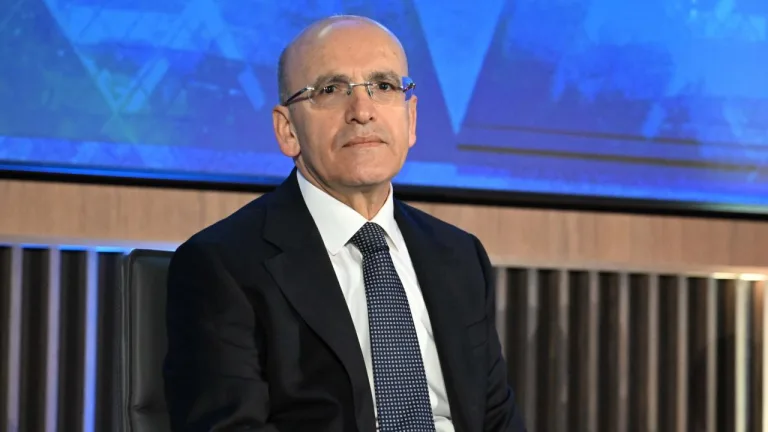 Bakan Şimşek konuşma yaparken kürsüde mikrofonlar önünde, arka planda ülke ve kurum simgeleri