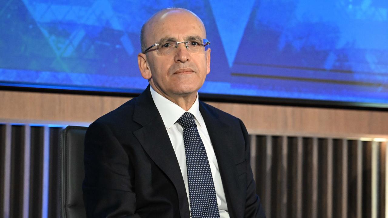 Bakan Şimşek konuşma yaparken kürsüde mikrofonlar önünde, arka planda ülke ve kurum simgeleri