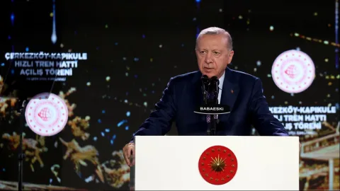 Erdoğan konuşma yaparken kürsüde mikrofonlara yönelmiş, arka planda Türk bayrağı ve kalabalık görünüyor