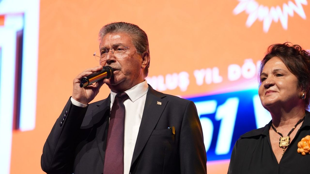 Üstel konuşurken el hareketi yapıyor; arka planda nötr bir zemin ve mikrofona yakın görünüm