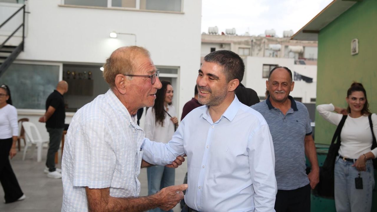 Aktunç konuşma yaparken kalabalığa bakıyor; arka planda belediye logosu ve bayraklar görünüyor